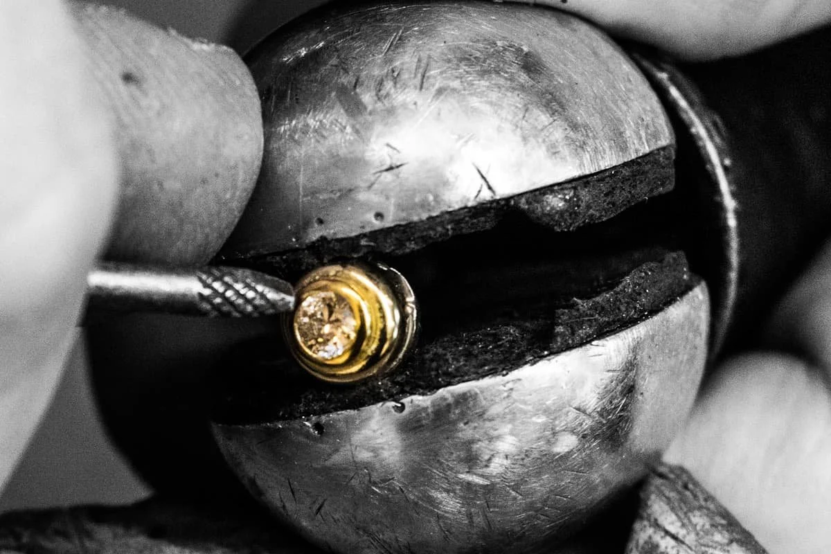 A jeweler's hand holds a diamond with a gold setting while working on a silver watch case in black and white.