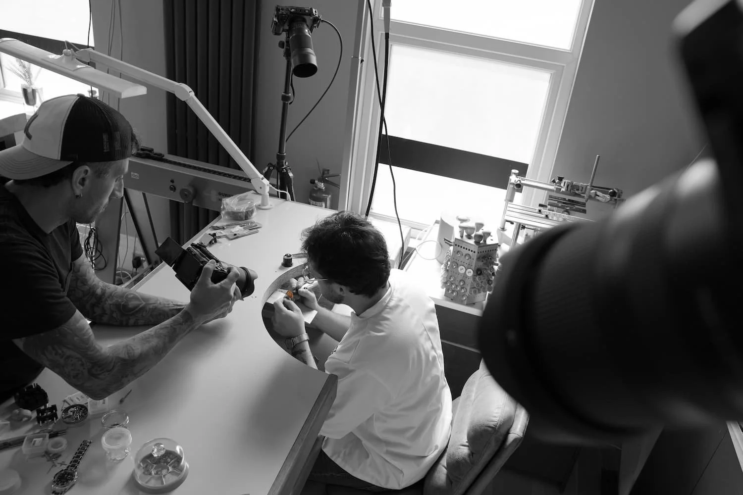 Two men working on a watch repair, one holding a camera, in a workspace with tools and equipment, near a window.