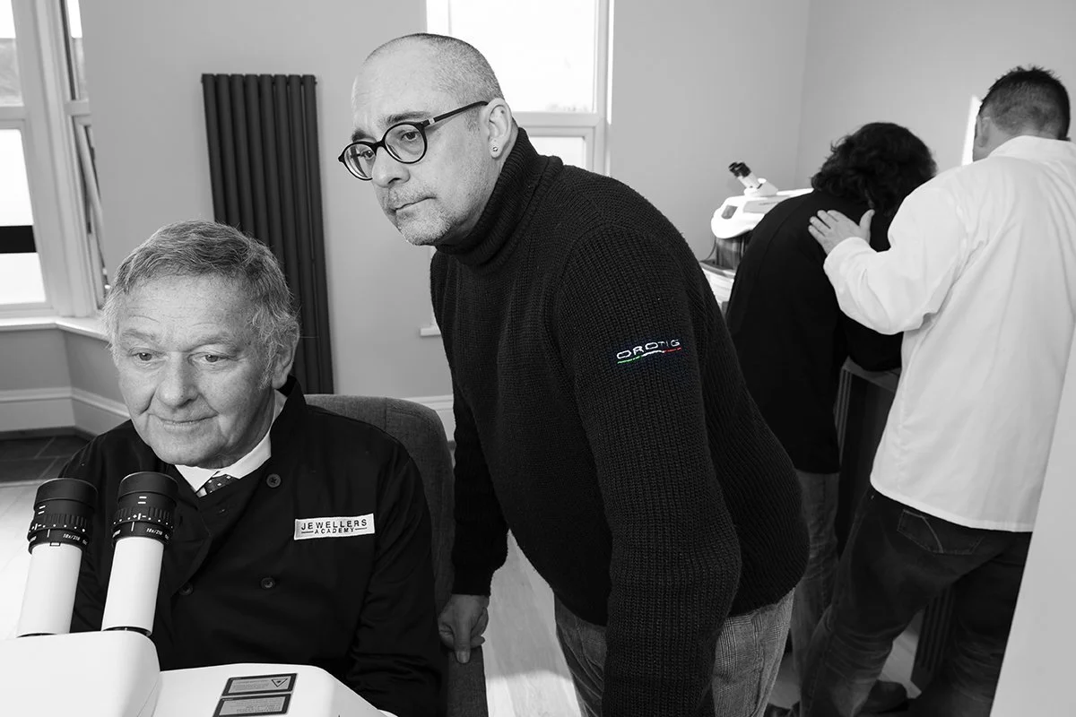 A man with glasses and a bald head leaning over a seated older man in a lab coat, with microscopes on a table, in a room with windows and a radiator.