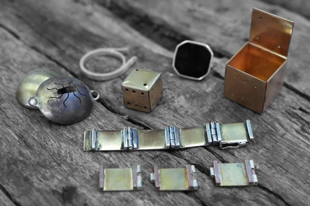 Various metal objects, including a small box with an open lid, a lock, a bent strip of metal, a dice, and a small rectangular container, are arranged on a weathered wooden surface.