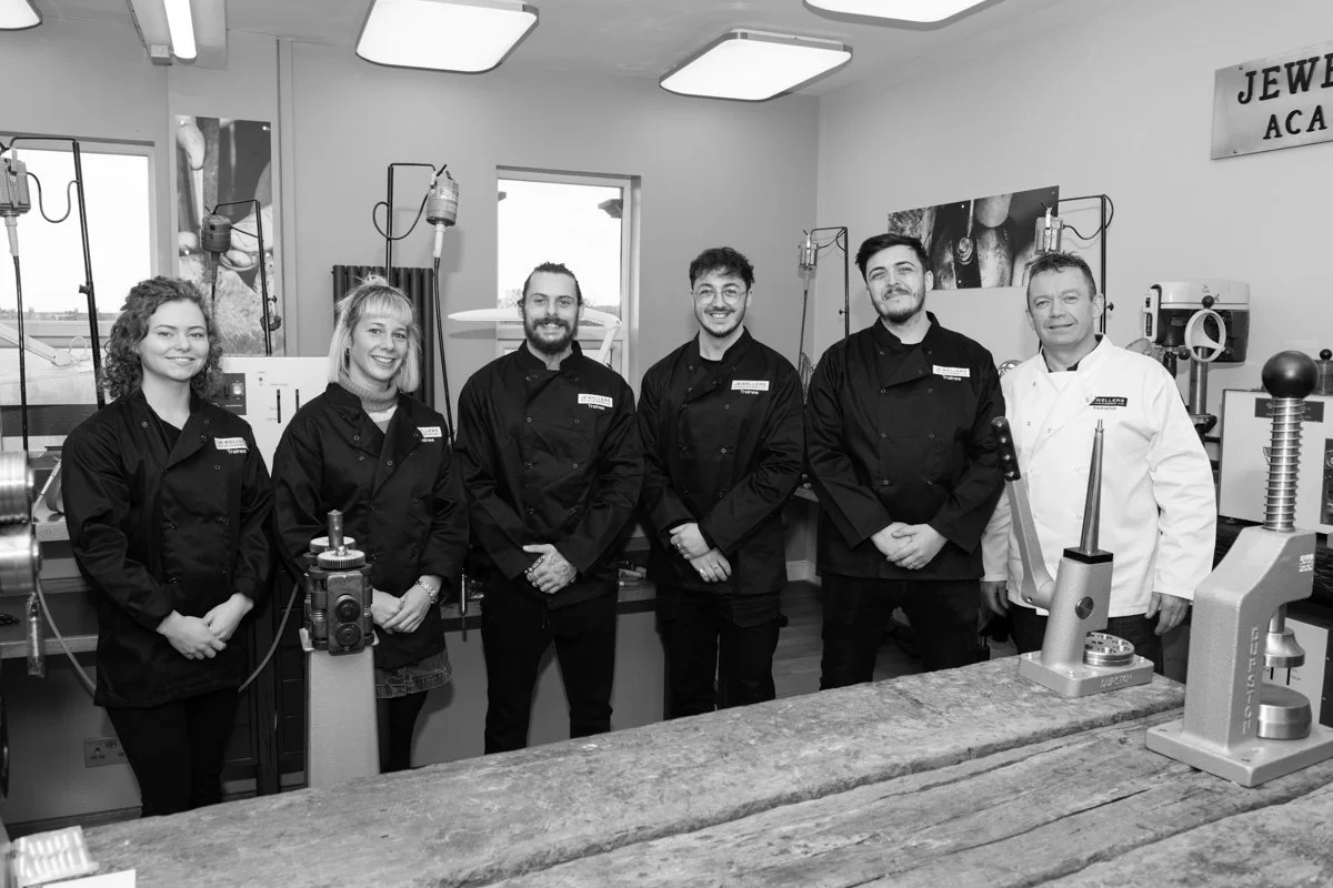 Group of six people in a jewelry workshop, wearing black uniforms, standing behind a workbench with jewelry-making tools and equipment, smiling at the camera.