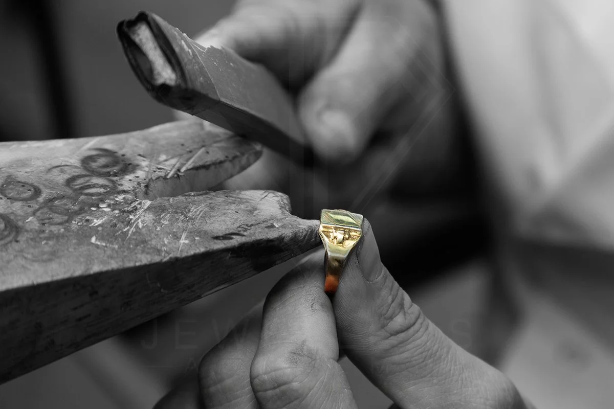A jeweler's hand holding a gold ring with a green and red design, working on it with a tool in a workshop.