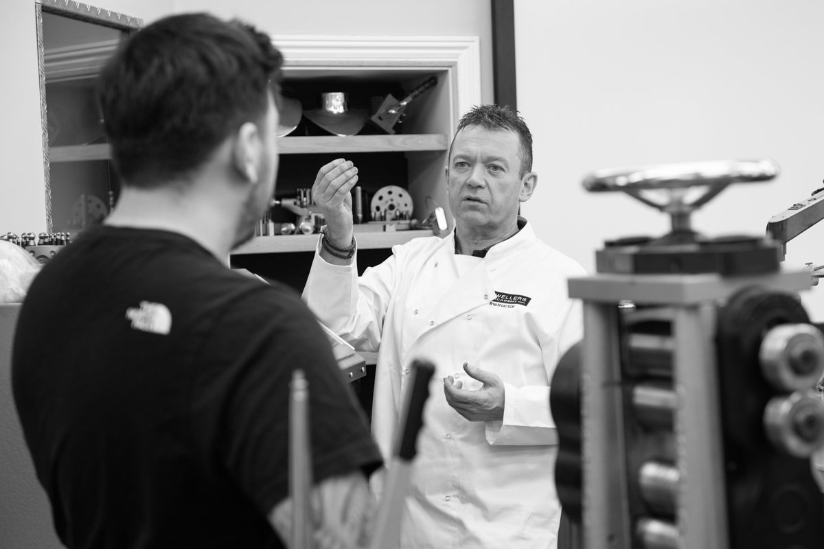 A man in a white lab coat explaining something to a younger man in a black T-shirt in a workshop with machinery and tools.