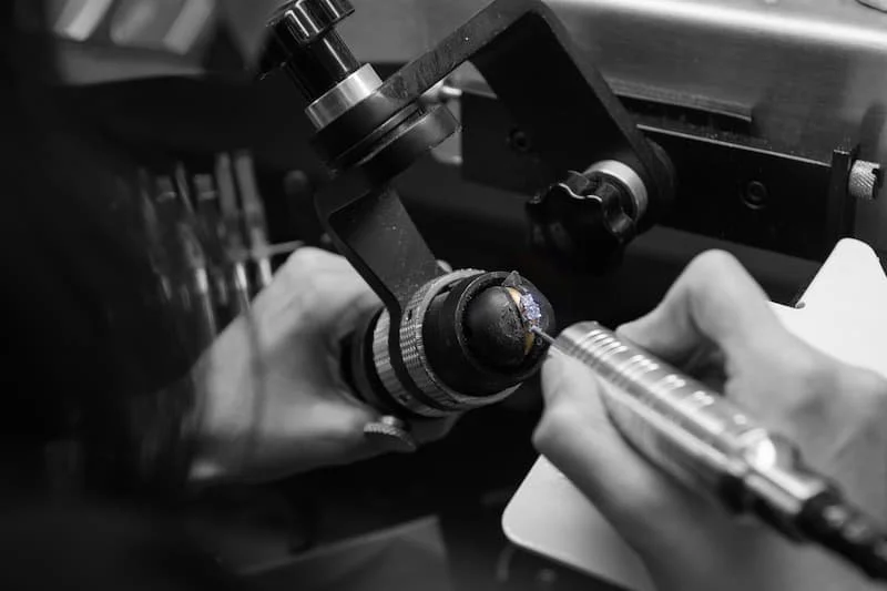 Close-up of a scientist or technician using a microscope to examine a small object, with a hand holding a tool or instrument near the microscope.