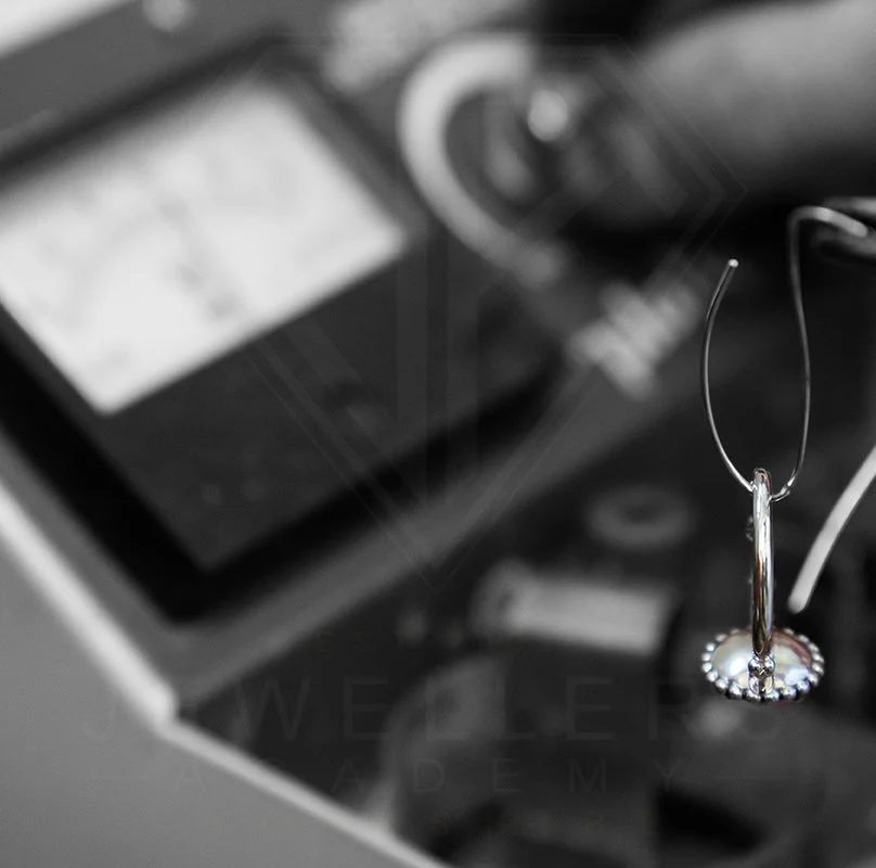 A pair of earrings hanging from a metal stand, with a blurred background showing a calculator and some other objects.