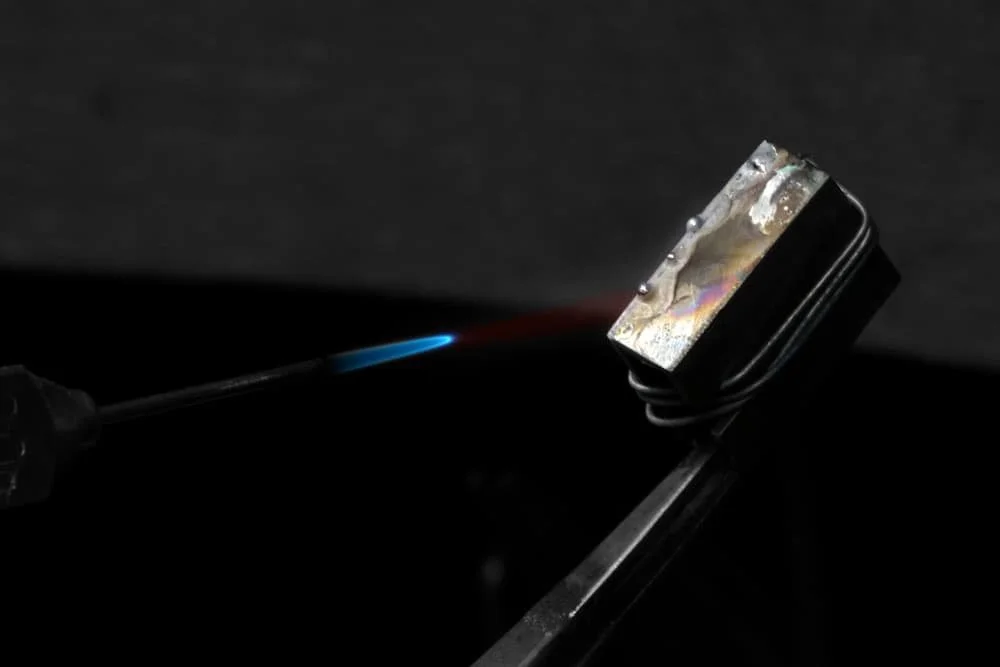 A soldering iron with a blue flame soldering a metal component in a dark setting.