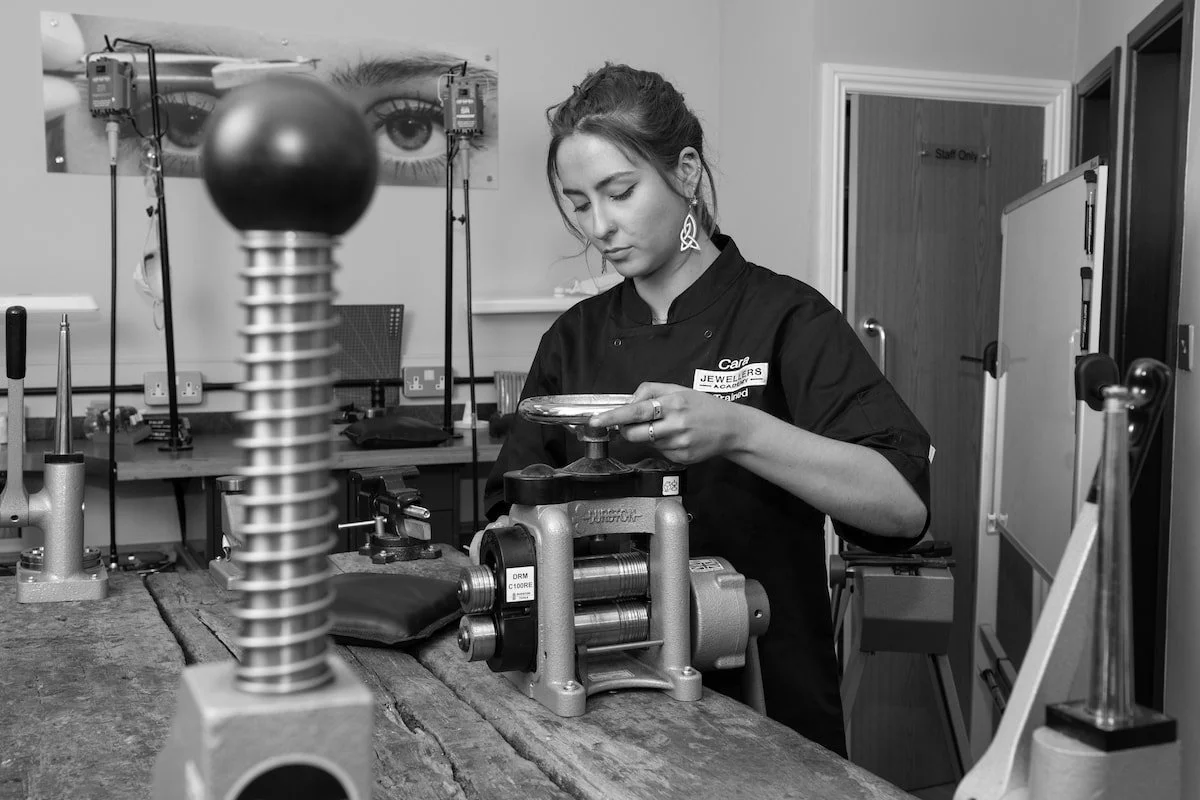 A woman working at a jewelry workshop, examining a piece with tools and equipment around her.