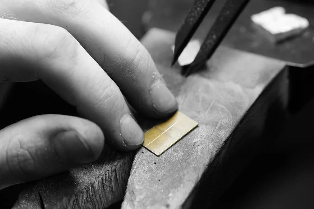 Close-up of a person's hand holding a small thin piece of gold sheet with tweezers, working on a craft project on a work surface.