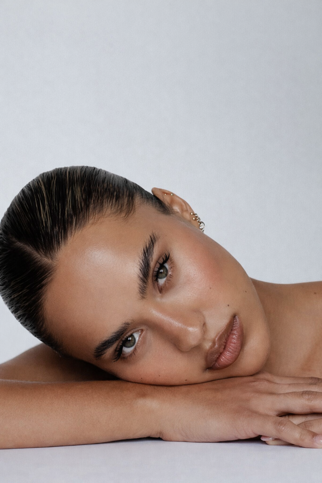 Portrait d'une femme aux cheveux bruns, maquillage naturel, repose sa tête sur son bras, fond blanc.