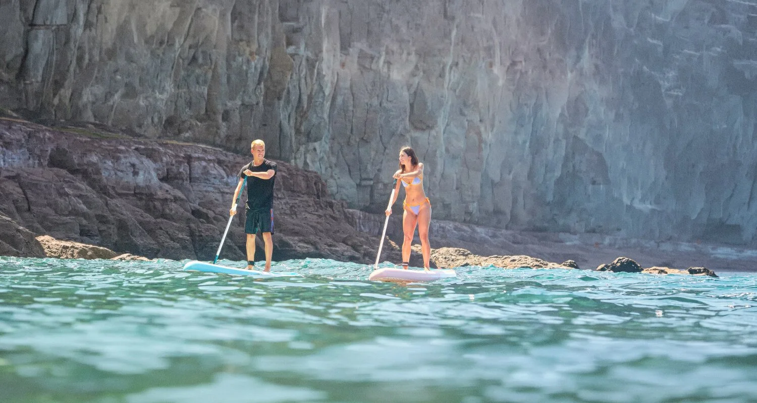 Deux personnes, un homme et une femme, font du paddle sur la mer près d'une falaise rocheuse.