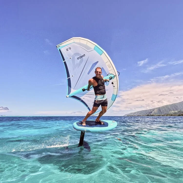 Homme pratiquant le flyboard avec une voile au dessus de l'eau claire et turquoise, paysage côtier en arrière-plan, ciel ensoleillé.