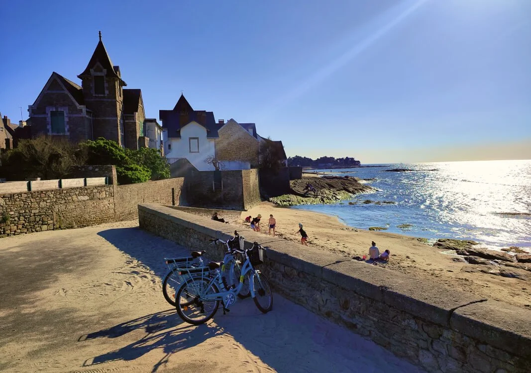 Plage avec des familles, deux vélos, bâtiments historiques en pierre sur la côte, soleil brillant sur la mer.