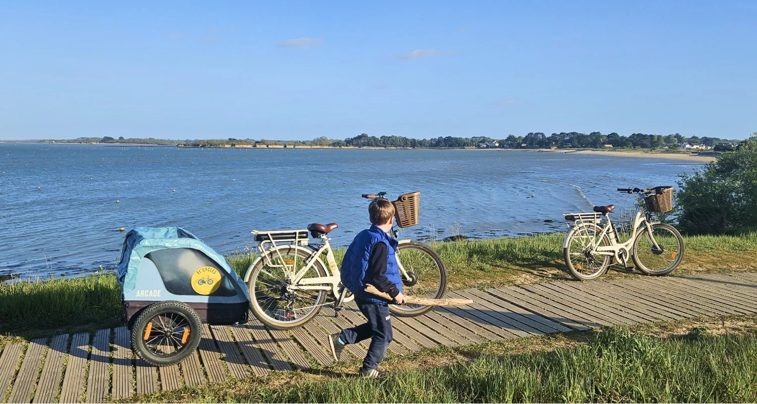 Un garçon avec un sac à dos qui pousse une remorque attachée à une bicyclette près d'un lac, avec une autre bicyclette stationnée sur le chemin en bois à côté de lui.