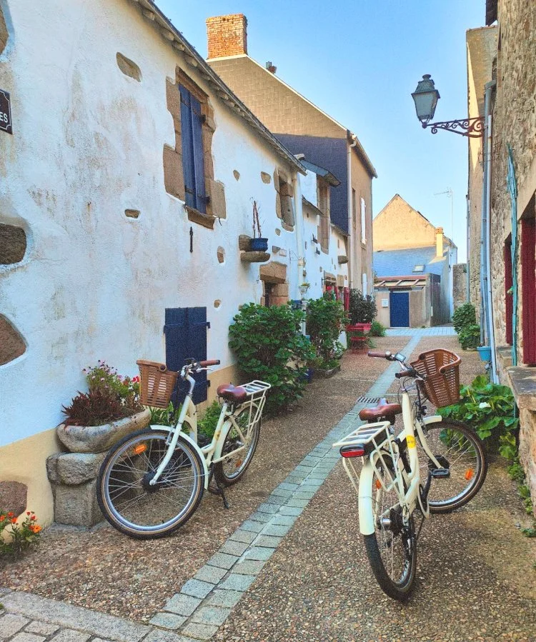 Ruelle pavée bordée de maisons anciennes avec deux vélos stationnés, quelques plantes en pot et un lampadaire.