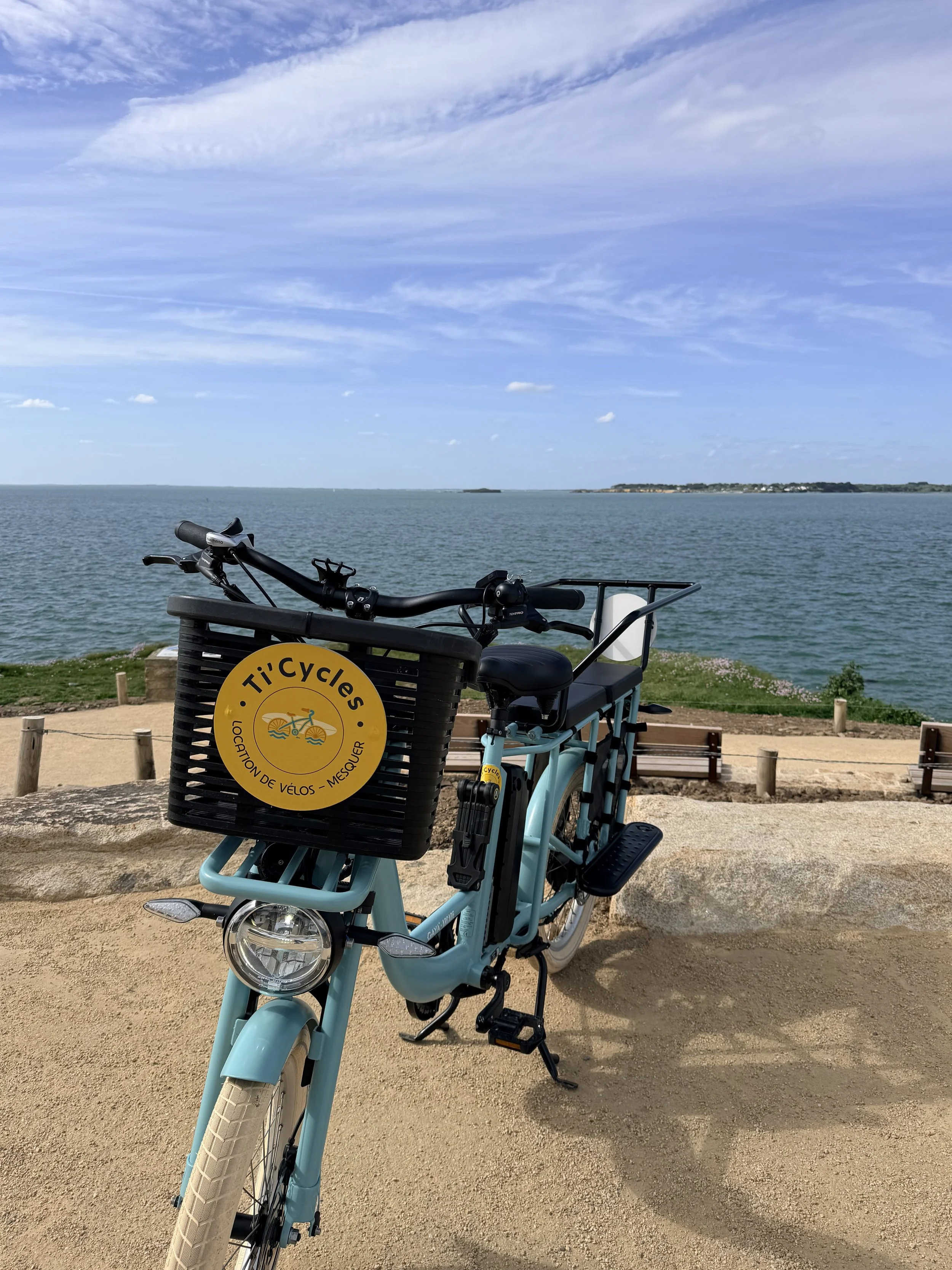 Location de vélo cargo électrique Ti’Cycles avec panier jaune, surplombant le littoral de Mesquer face à l'océan Atlantique par temps clair.