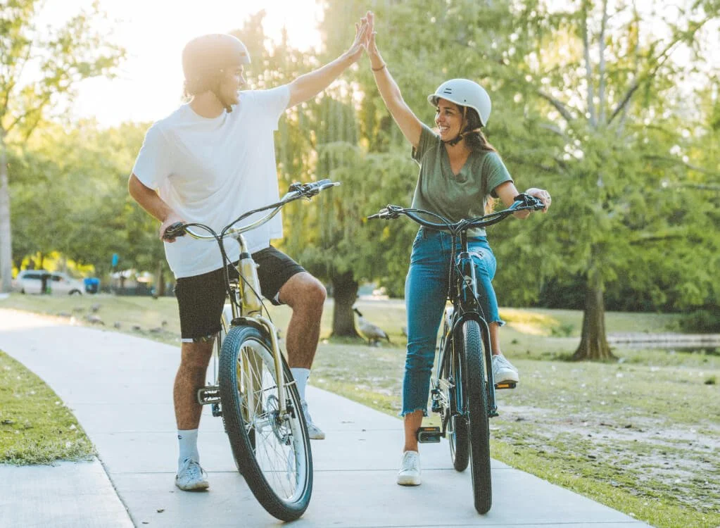 Deux personnes font du vélo sur un sentier dans un parc, elles se font un high-five, il fait soleil et il y a des arbres verts autour.