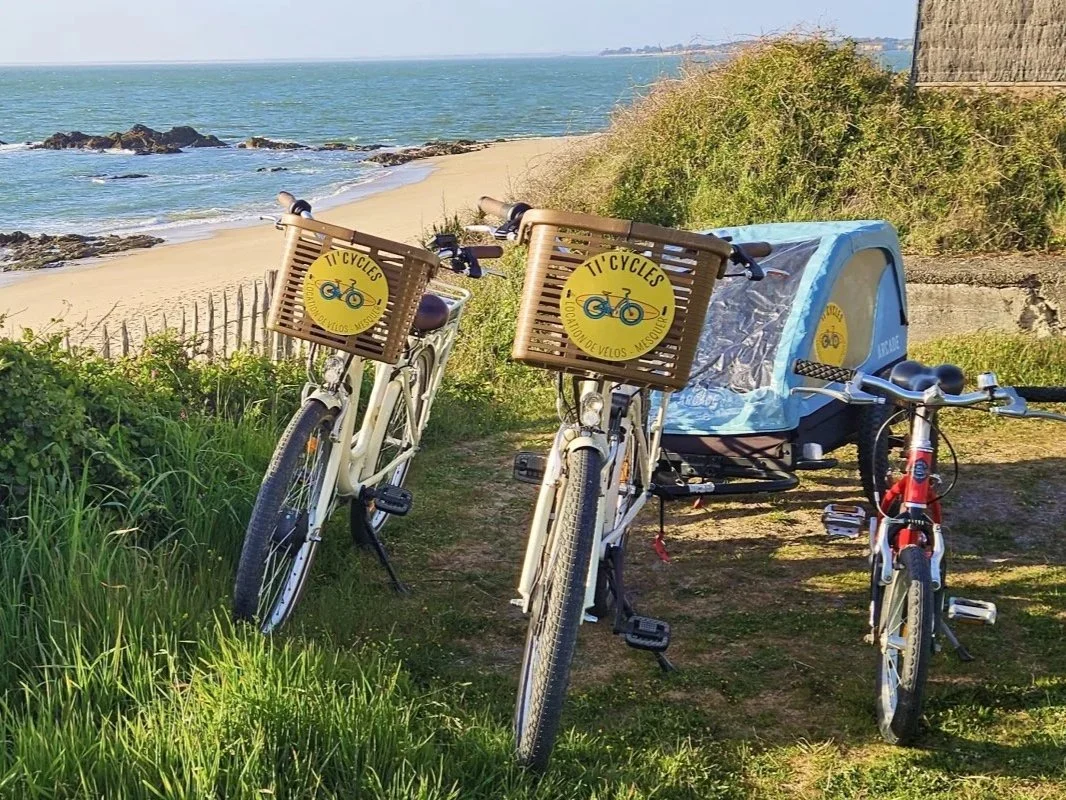 Deux vélos avec des paniers en bois portant un logo de TI Cycles sont garés sur un chemin près d'une plage avec des rochers, du sable et la mer en arrière-plan.