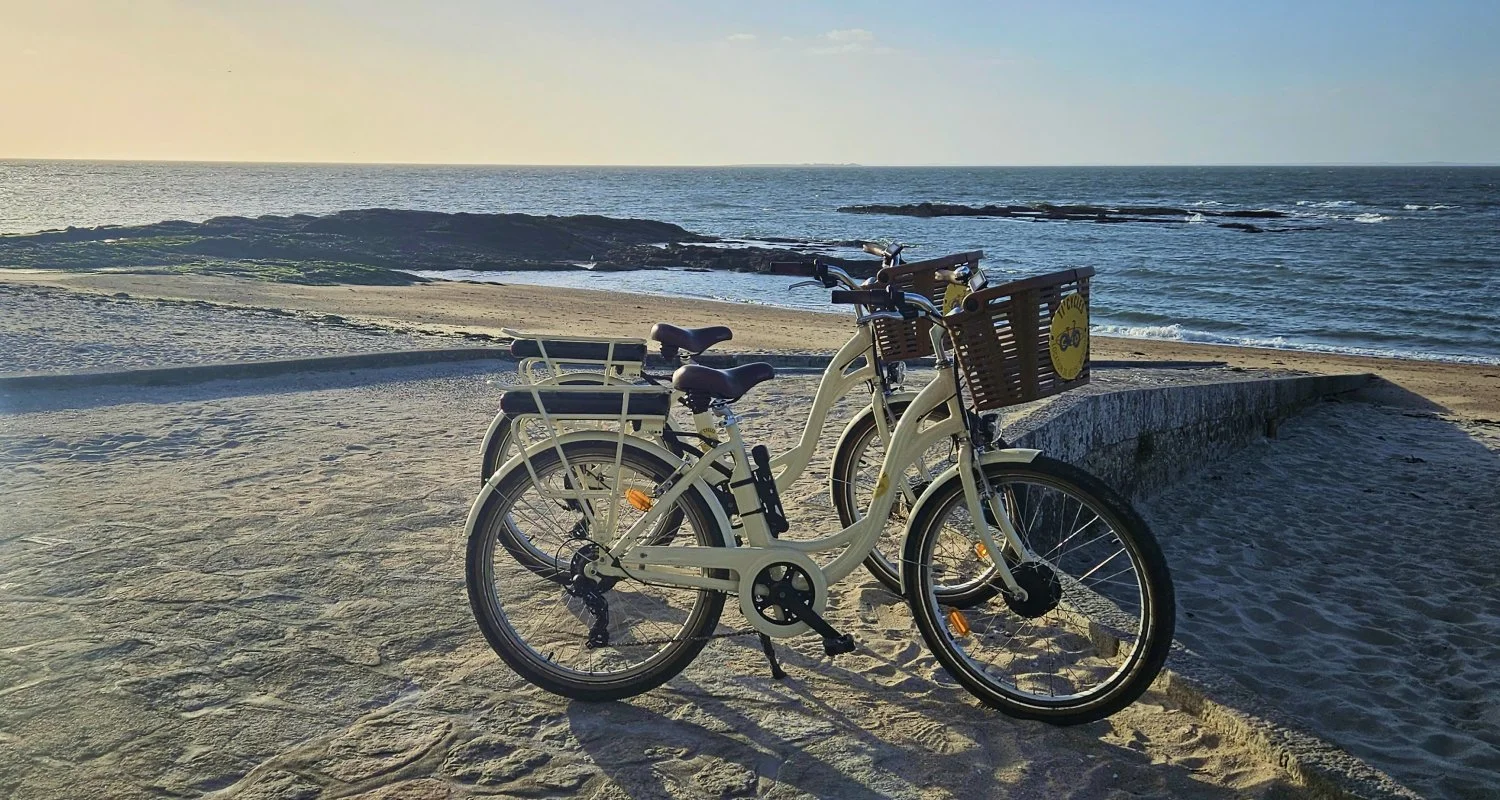 Deux vélos électriques avec paniers sur une plage au coucher du soleil, face à la mer.