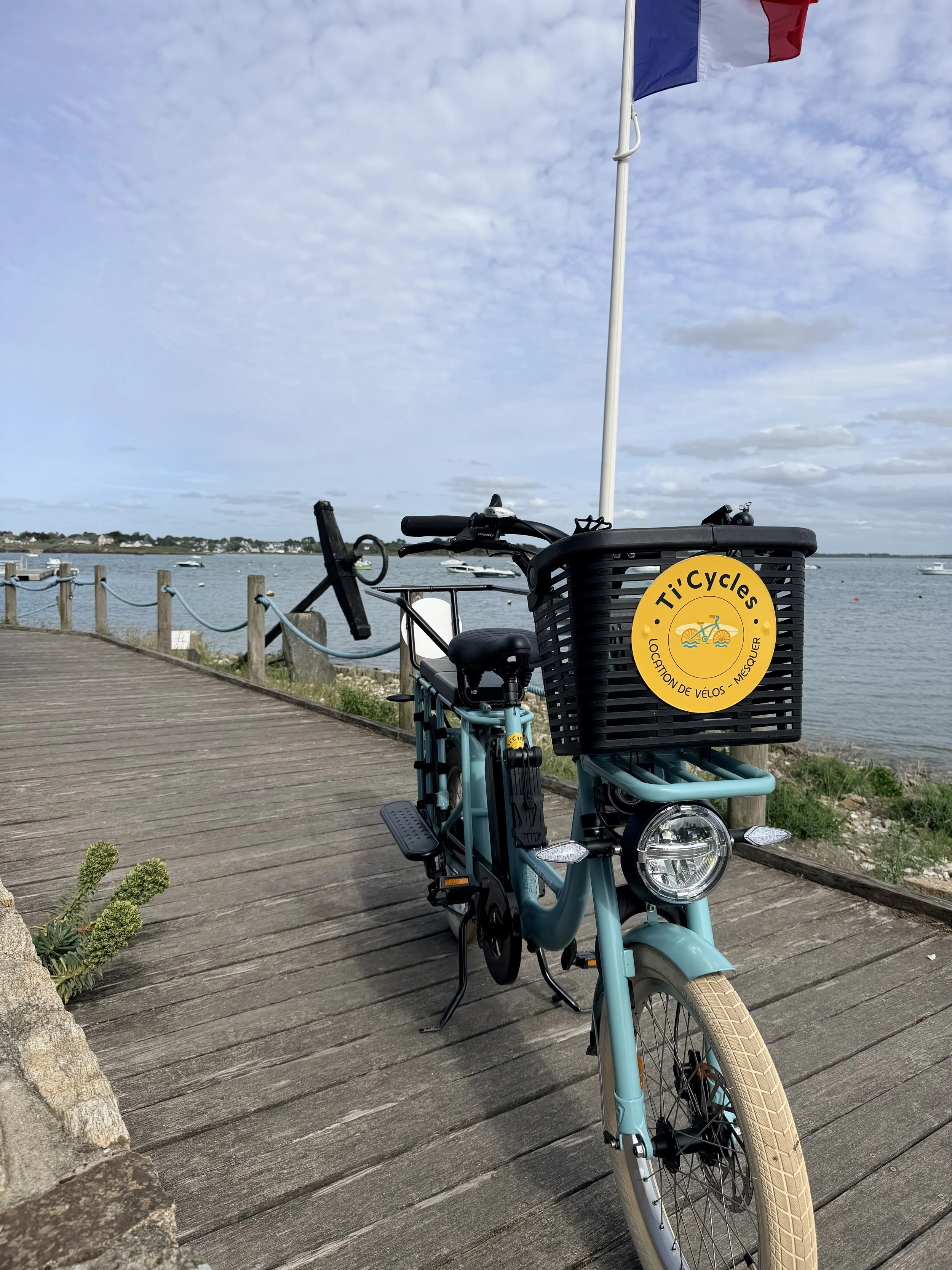 Vélo électrique longtail bleu de Ti’Cycles stationné sur un ponton en bois à Mesquer, avec panier avant, drapeau français et vue sur les bateaux en mer.