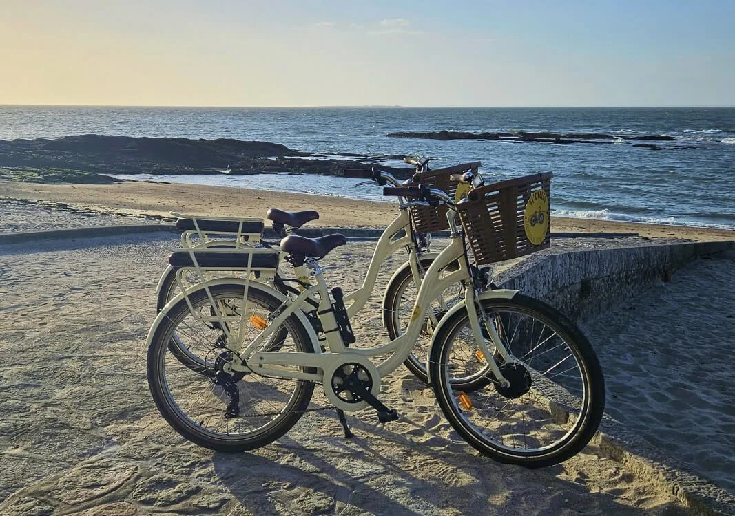 Deux vélos électriques blancs avec paniers en bois, stationnés sur la plage au coucher du soleil, face à la mer.