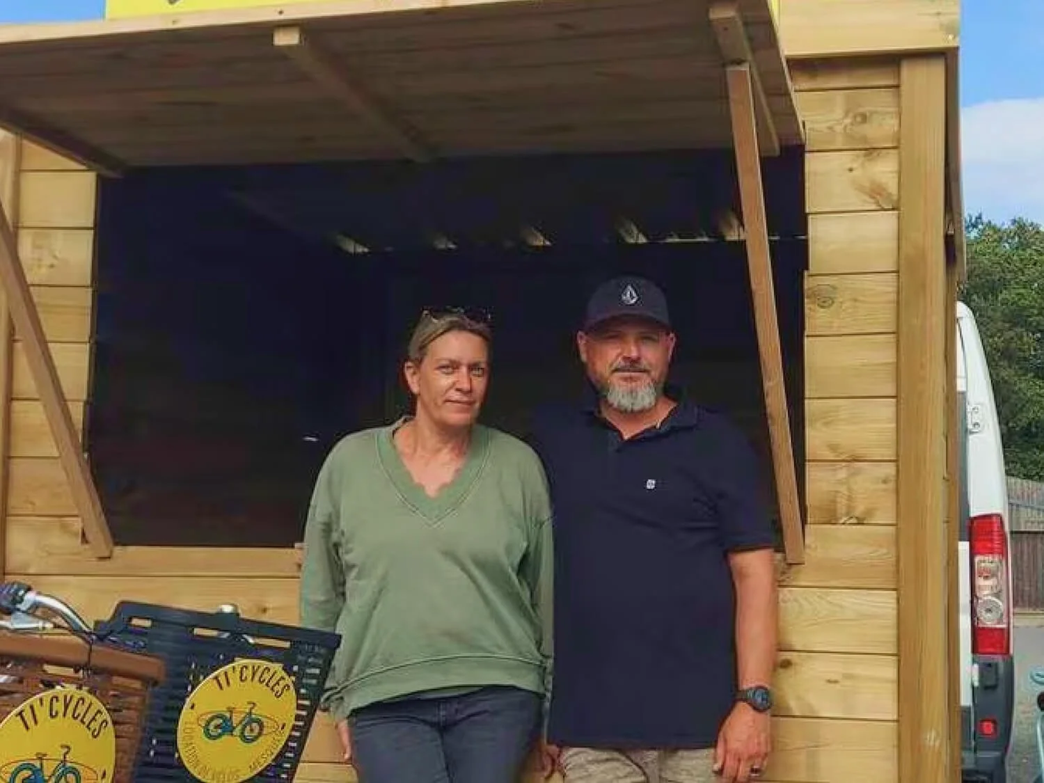 Deux personnes debout devant un stand en bois, une femme et un homme, avec des vélos et un signe 'Ti' Cycles' à côté.