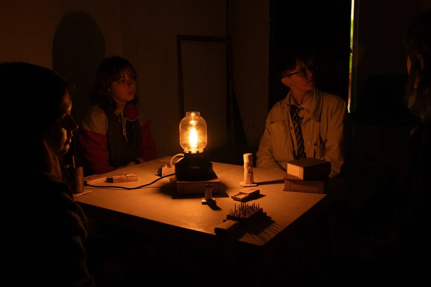 Four people sit around a table in a dark room, illuminated by a single vintage lantern, with books and small objects on the table.