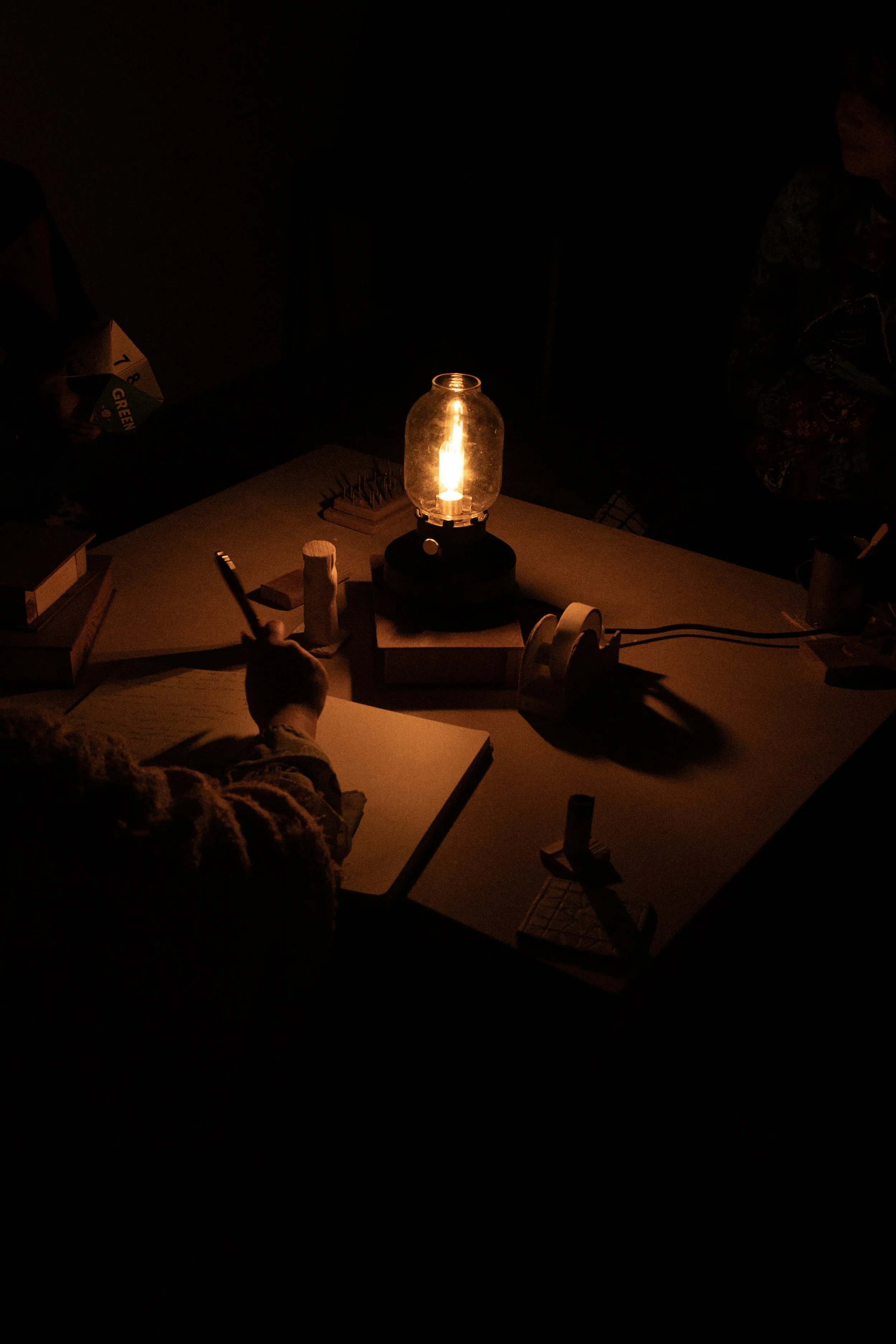 A dimly lit room with a table illuminated by a lantern, and people working or studying with notebooks and pens around the table.