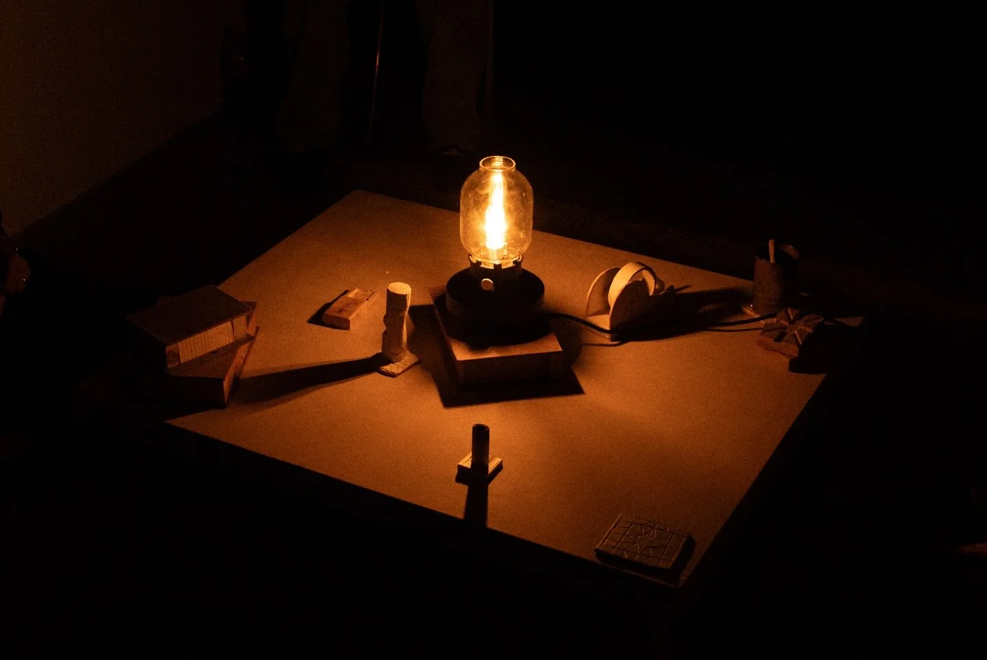 A table illuminated by a vintage oil lantern surrounded by various objects, including matches, a small container, and some paper machines, in a dark room.
