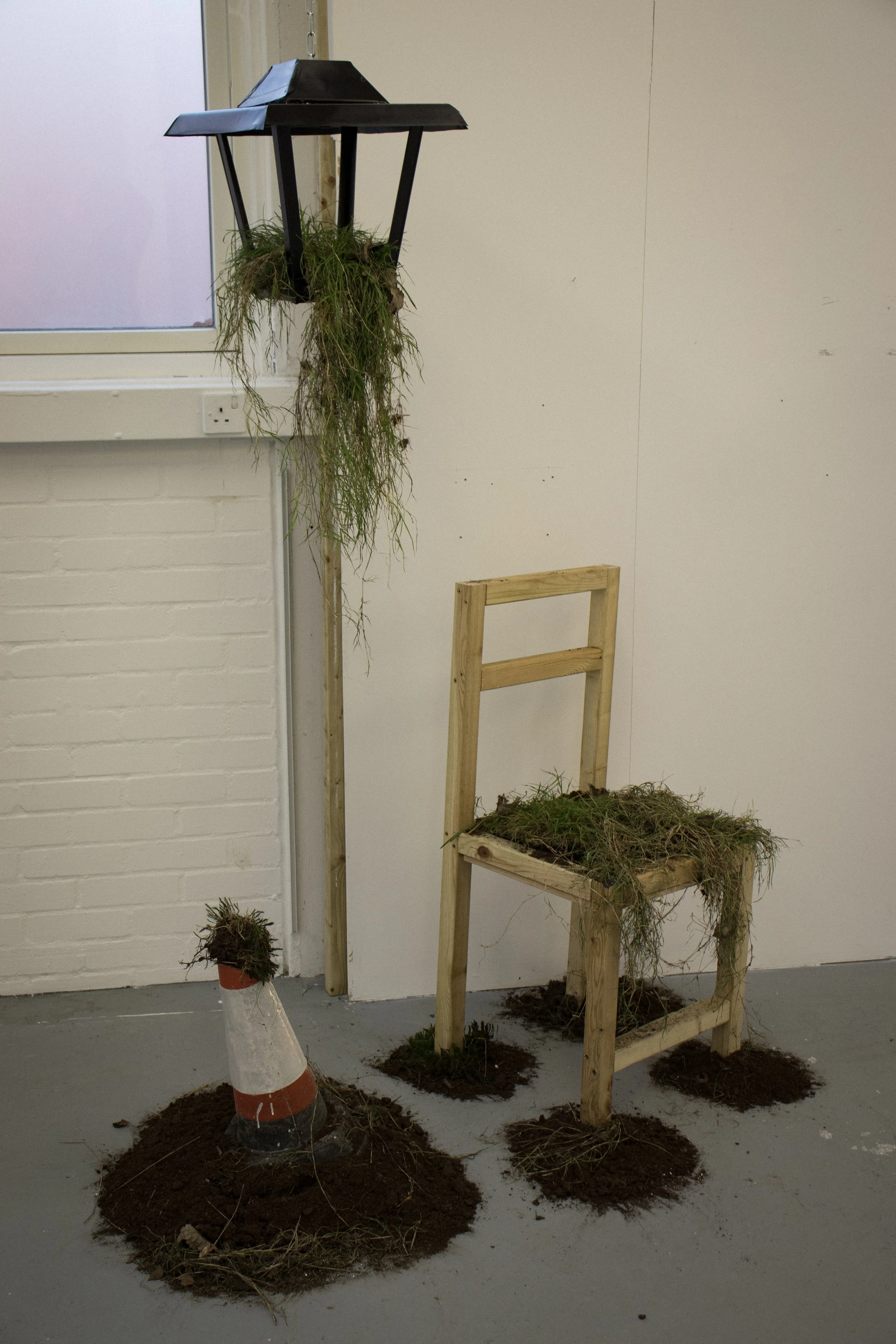 An art installation featuring a black streetlamp with hanging grass, a small potted grass plant, a wooden chair with grass on the seat, and soil mounds on the floor, in a room with a white brick wall and a window.