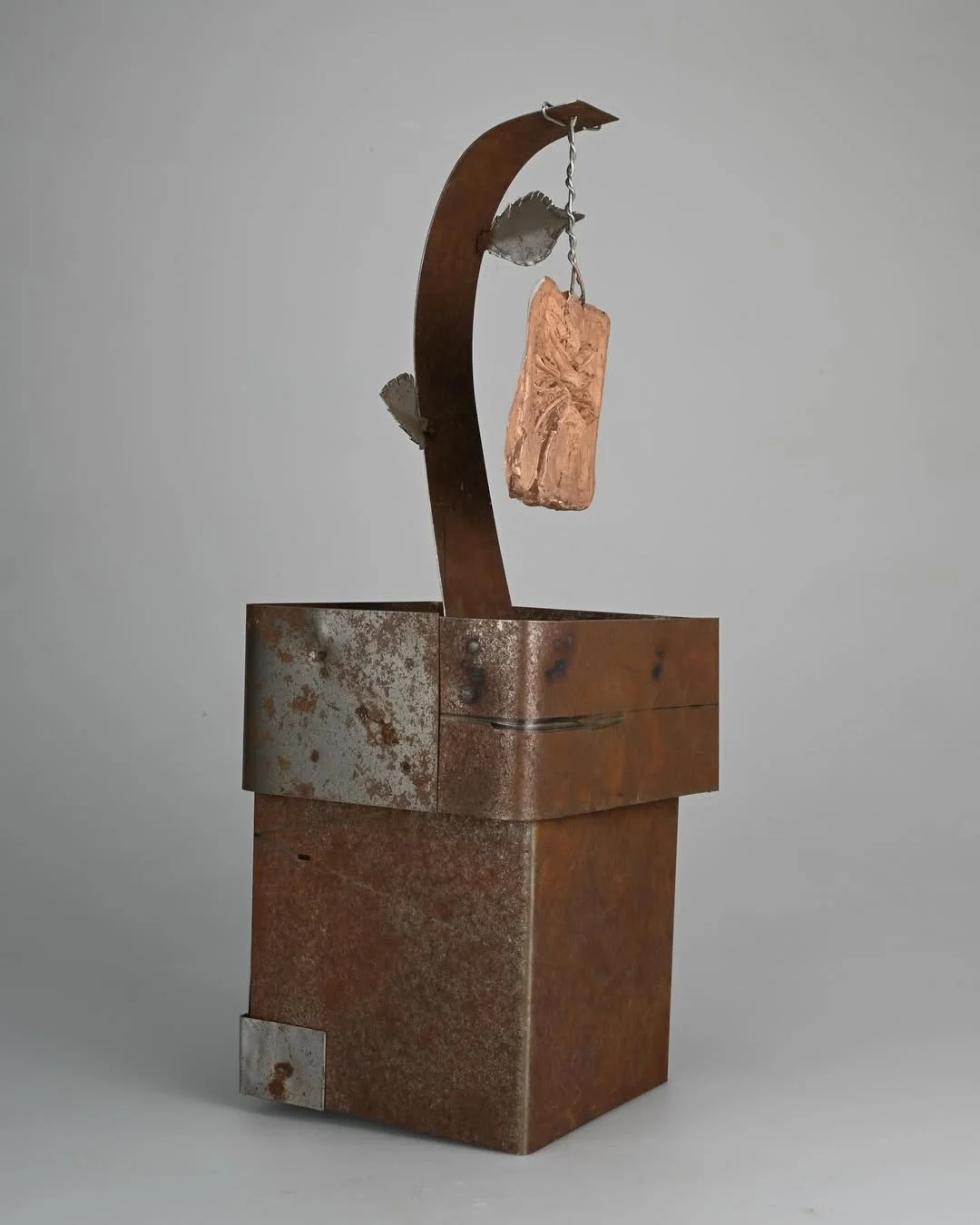 Rusty metal sculpture resembling a hanging brick, supported by a curved metal arch with leaf-like attachments, set against a plain gray background.