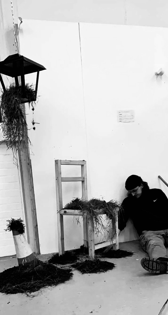 A person sitting on the floor next to a display with plants and wooden structures, including a small table with plants and a cone-shaped plant holder, in an indoor setting.