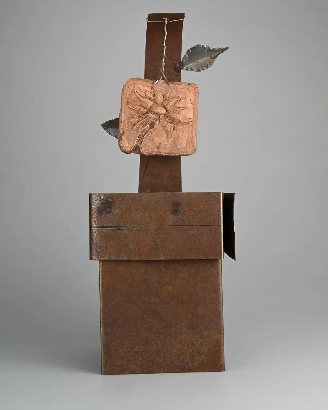 An abstract sculpture made of rusted metal pieces, featuring a rectangular block with a carved flower, a tall rectangular vertical piece, and a leaf-like element attached to the vertical piece.