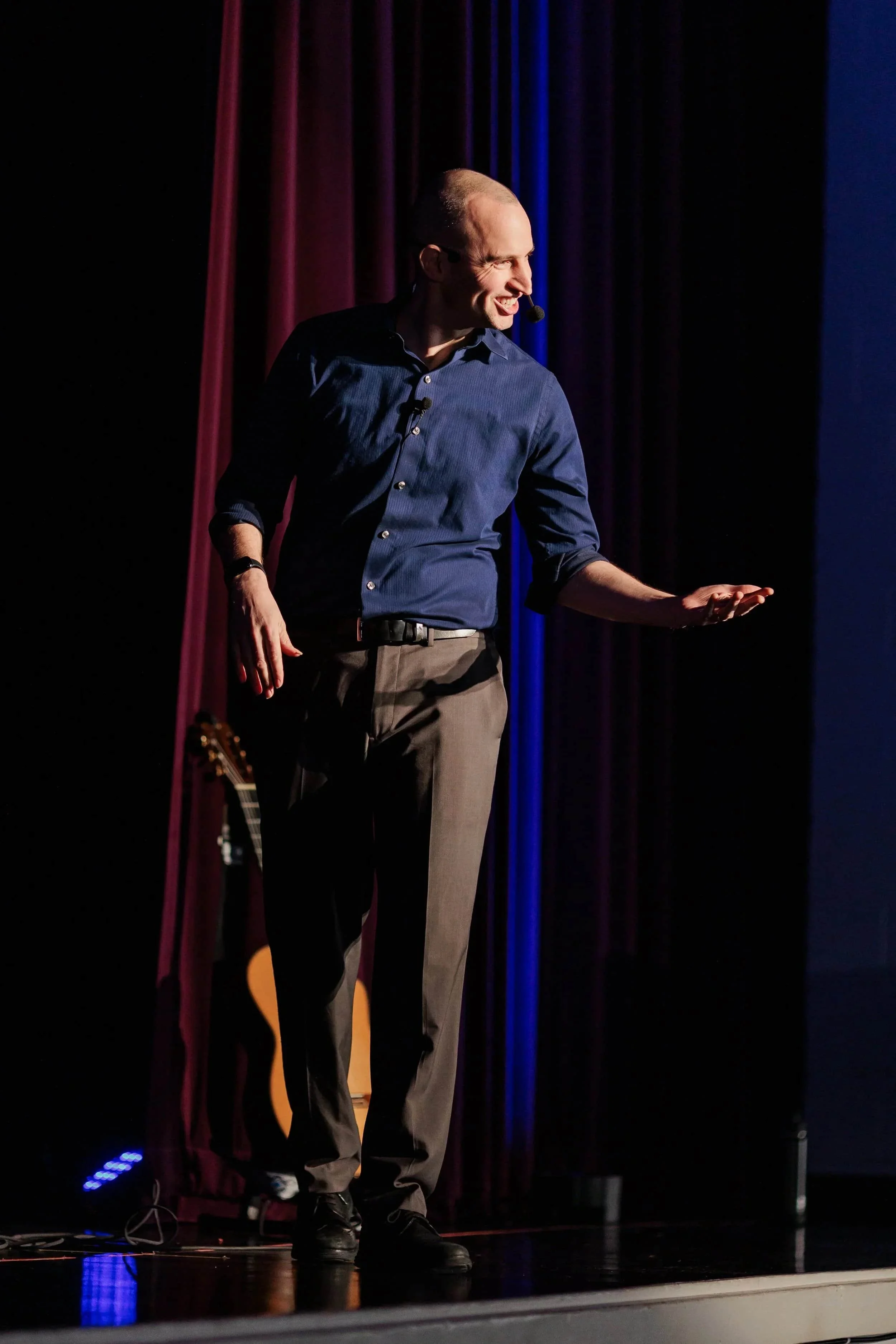 A man in a blue shirt speaks on stage with a headset microphone, gesturing with one hand. A guitar is visible behind him.