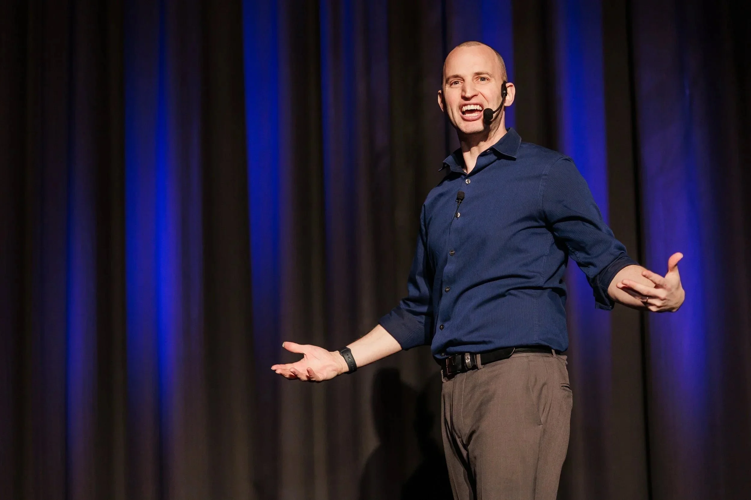 A man in a blue shirt and gray pants giving a presentation on stage with a dark curtain background.