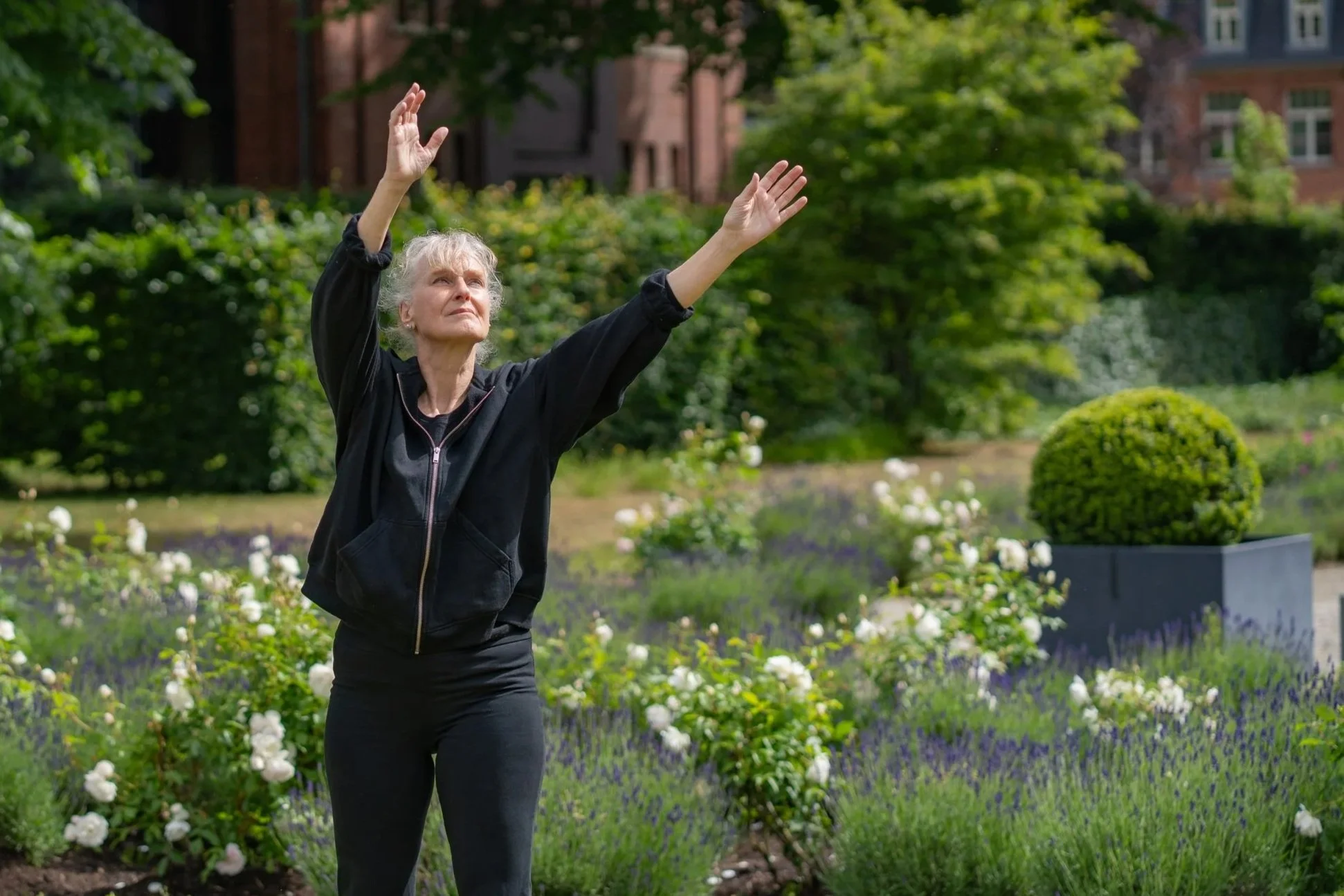 Silke Radtke macht eine Qigong Übung. Im Hintergund sind viele Grüne und Lila Blumen zu sehen.