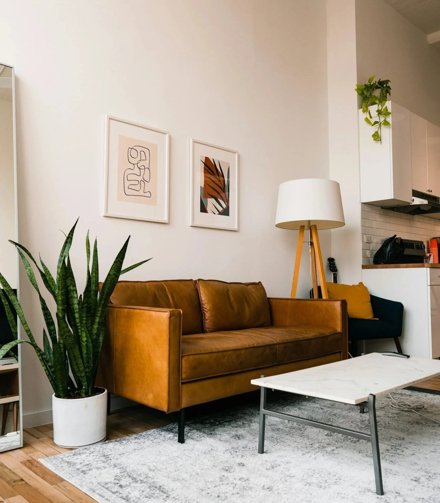 Living room with a brown leather sofa, a potted snake plant, a white marble coffee table, a floor lamp with a white shade and wooden tripod, wall art, and a small black chair with a yellow pillow in an open kitchen.