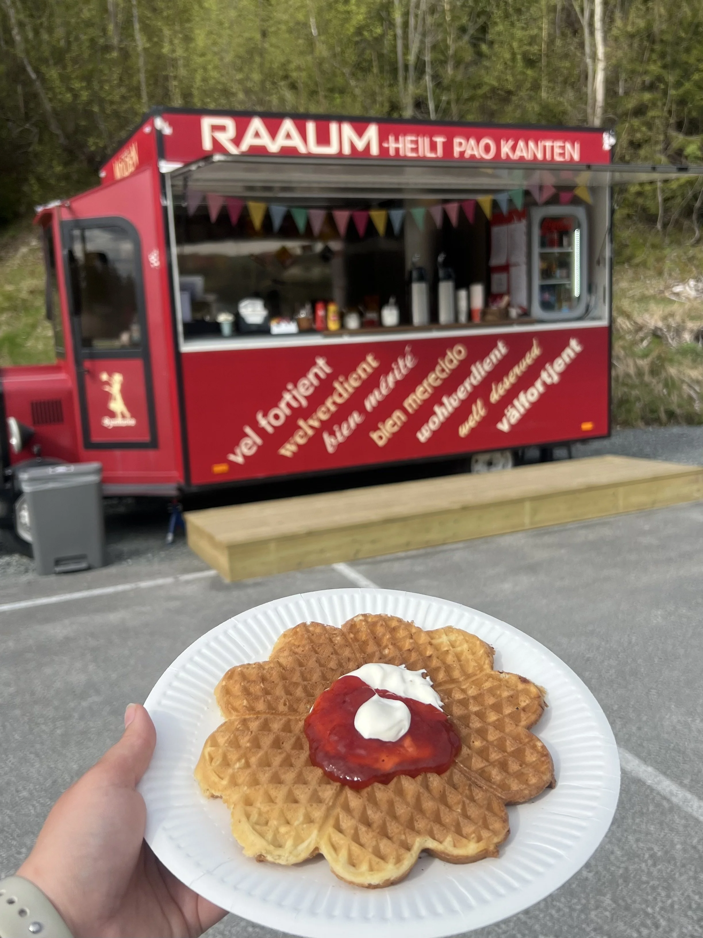 En person holder en tallerken med hvetewafler, toppet med jordbærsyltetøy og rømme, foran en matvogn med svensk tekst.