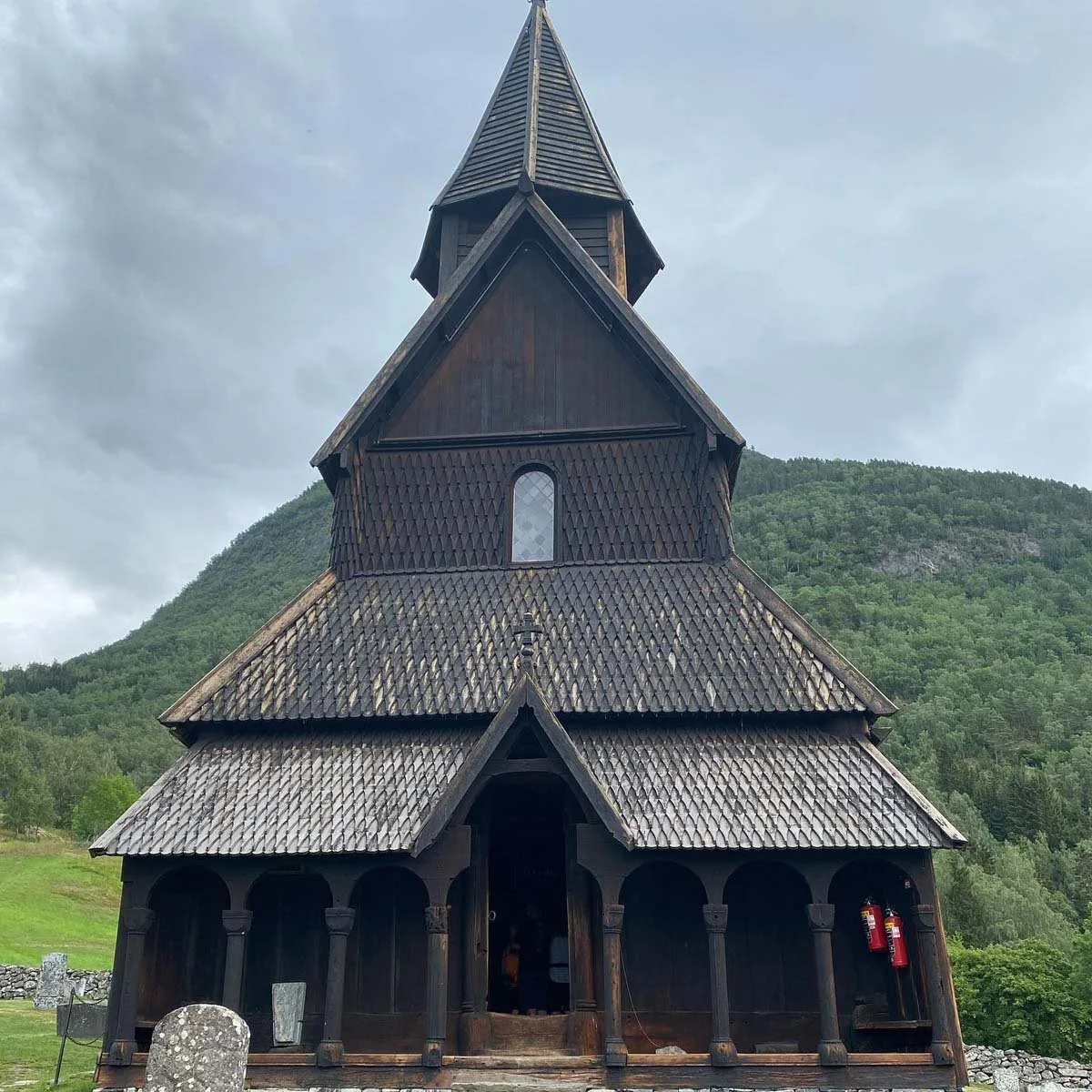 En svart trekirke med et høyt spisst tårn, omgitt av fjell og grønn natur under en overskyet himmel.