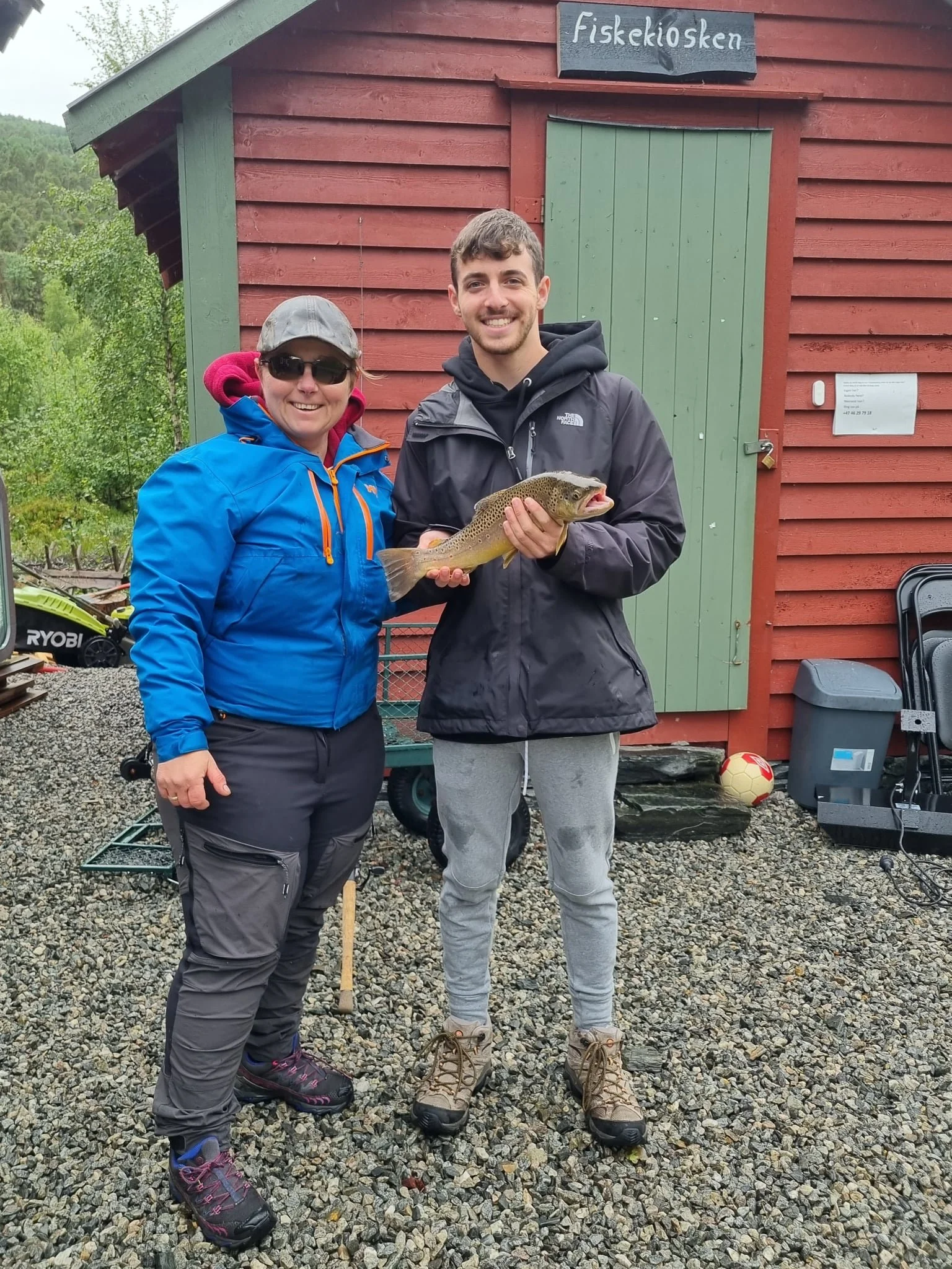 To personer står utenfor en rød liten fiskebod, en av dem holder en fisk. De smiler mot kamera.
