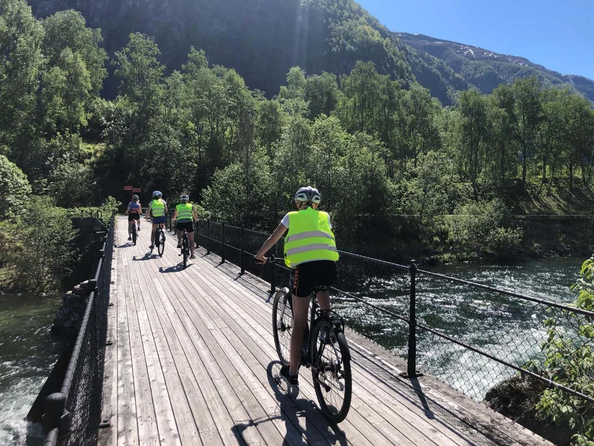 Fem personer sykler på en trebro over en elv, omgitt av grønne fjell og trær, på en solfylt dag.