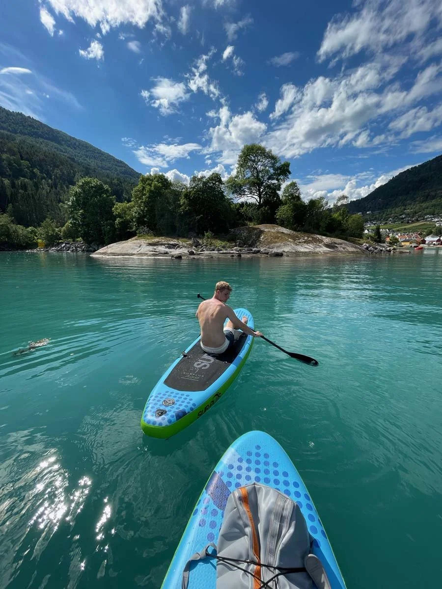 En person padler på en rolig, grønnfarget innsjø omgitt av grønne trær og fjell under en skyet, men solrik himmel, sett fra en annen persons perspektiv som sitter på en blå padlebre.