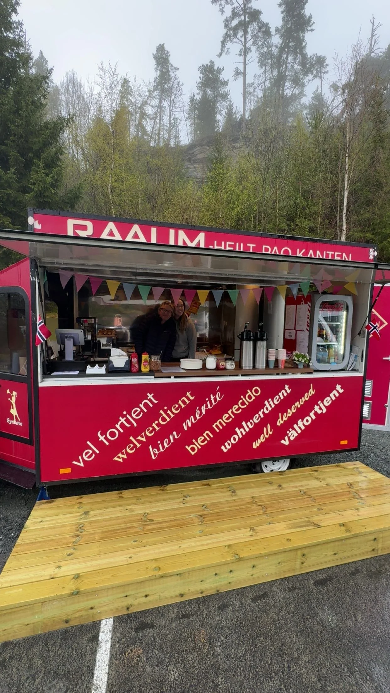 En rød matvogn med tekst på norsk og flere språk, som tilbyr mat og drikke, med to personer som står bak disken, omgitt av trær og en fjellveggen bak.