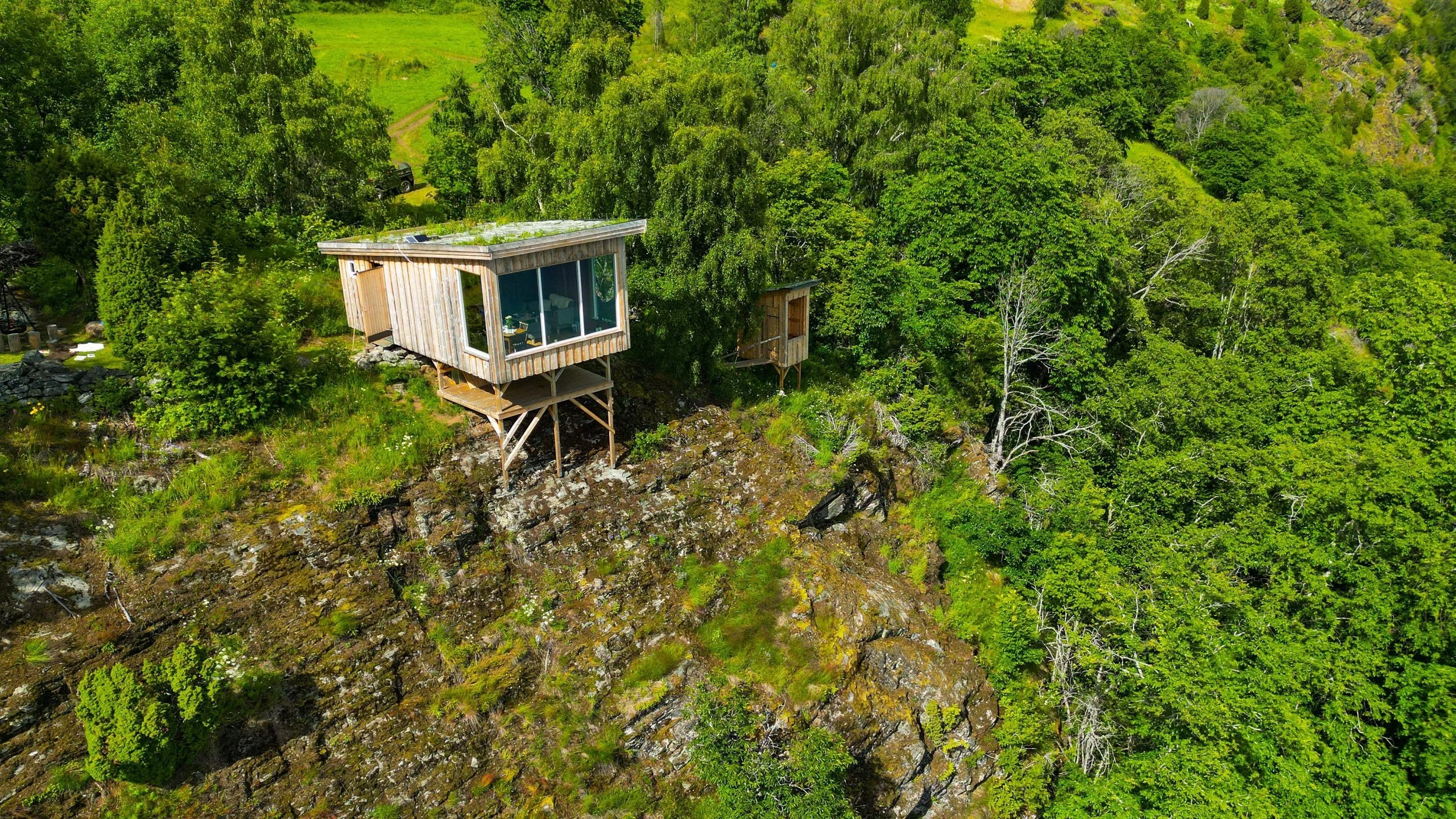Fjelltomt med en moderne trehytte omgitt av grønn vegetasjon og stein, sett fra luften