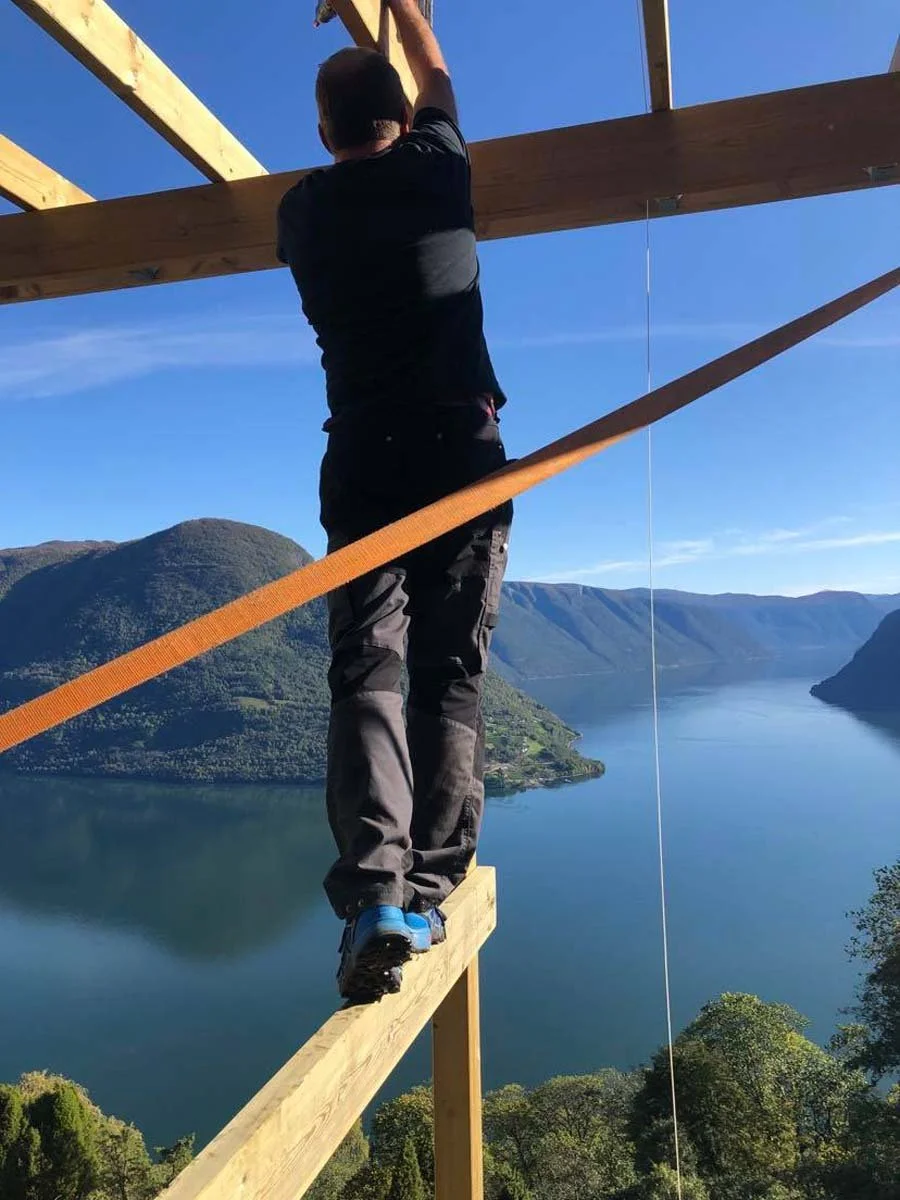 En person jobber på en stillas med utsikt over en innsjø omgitt av fjell, under en klar blå himmel.