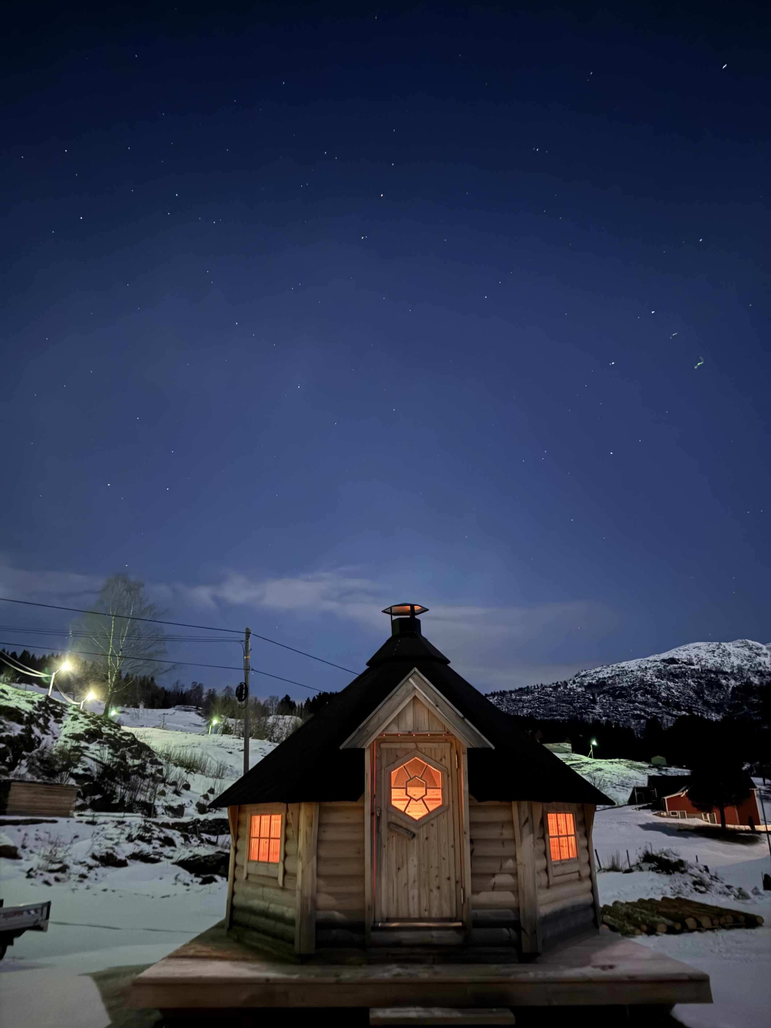 En liten trebygning med opplyste vinduer og dør, omgitt av snø, under en klar stjerneklar natt, med fjell i bakgrunnen.