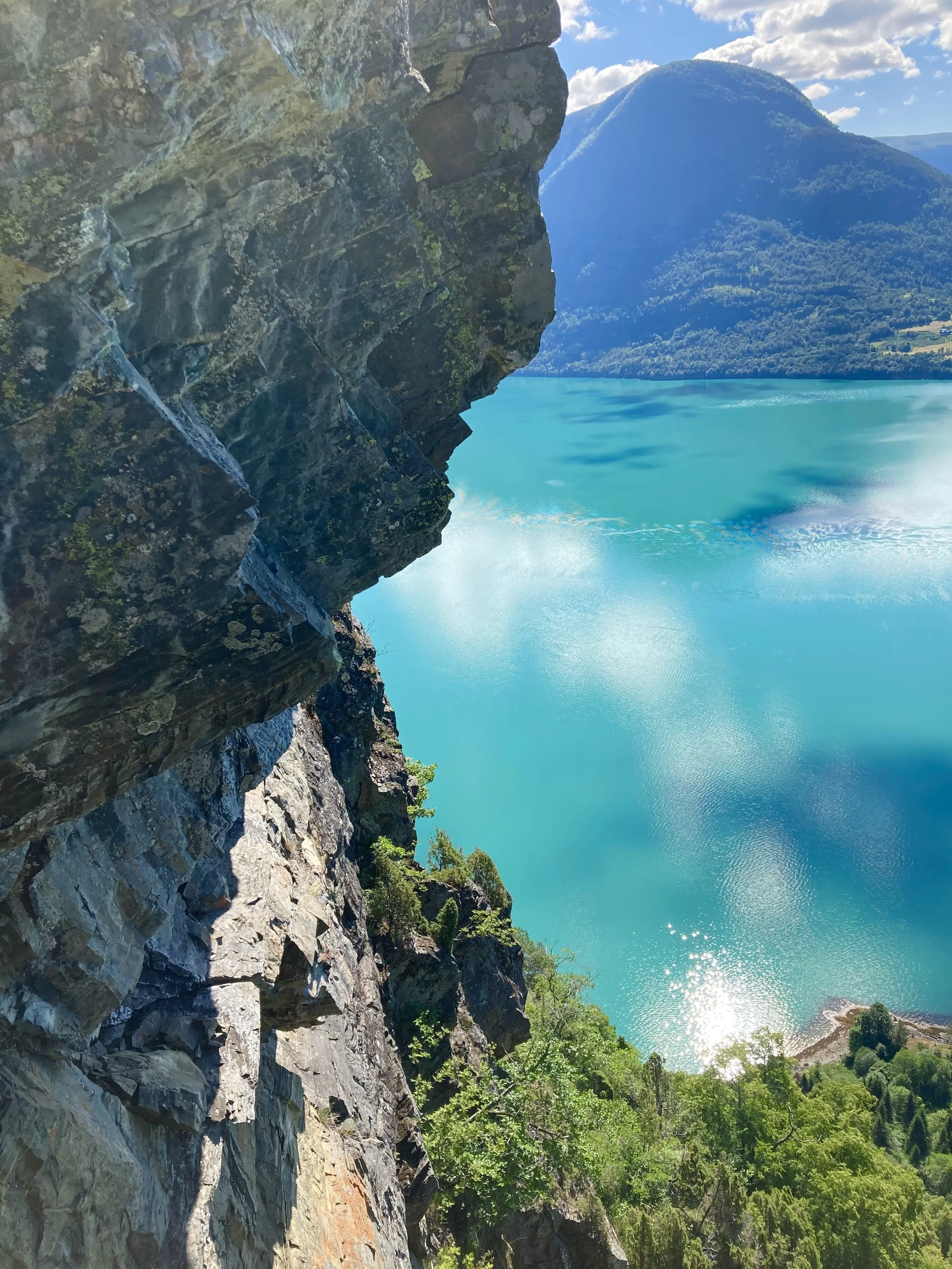 Utsikt fra en fjellvegg over en turkis innsjø omgitt av grønne skoger og fjell i bakgrunnen.