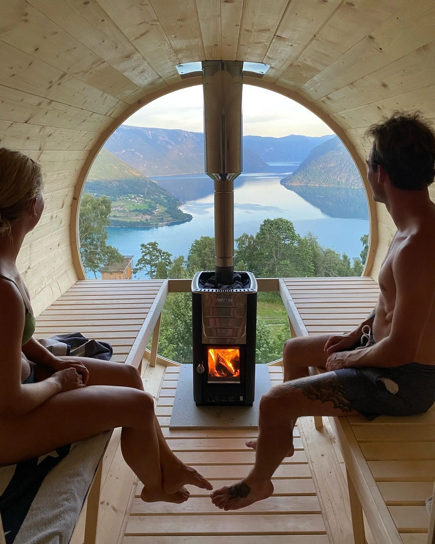 To personer sitter i en liten badstue med utsikt over en innsjø og fjell i bakgrunnen, ved en peis.