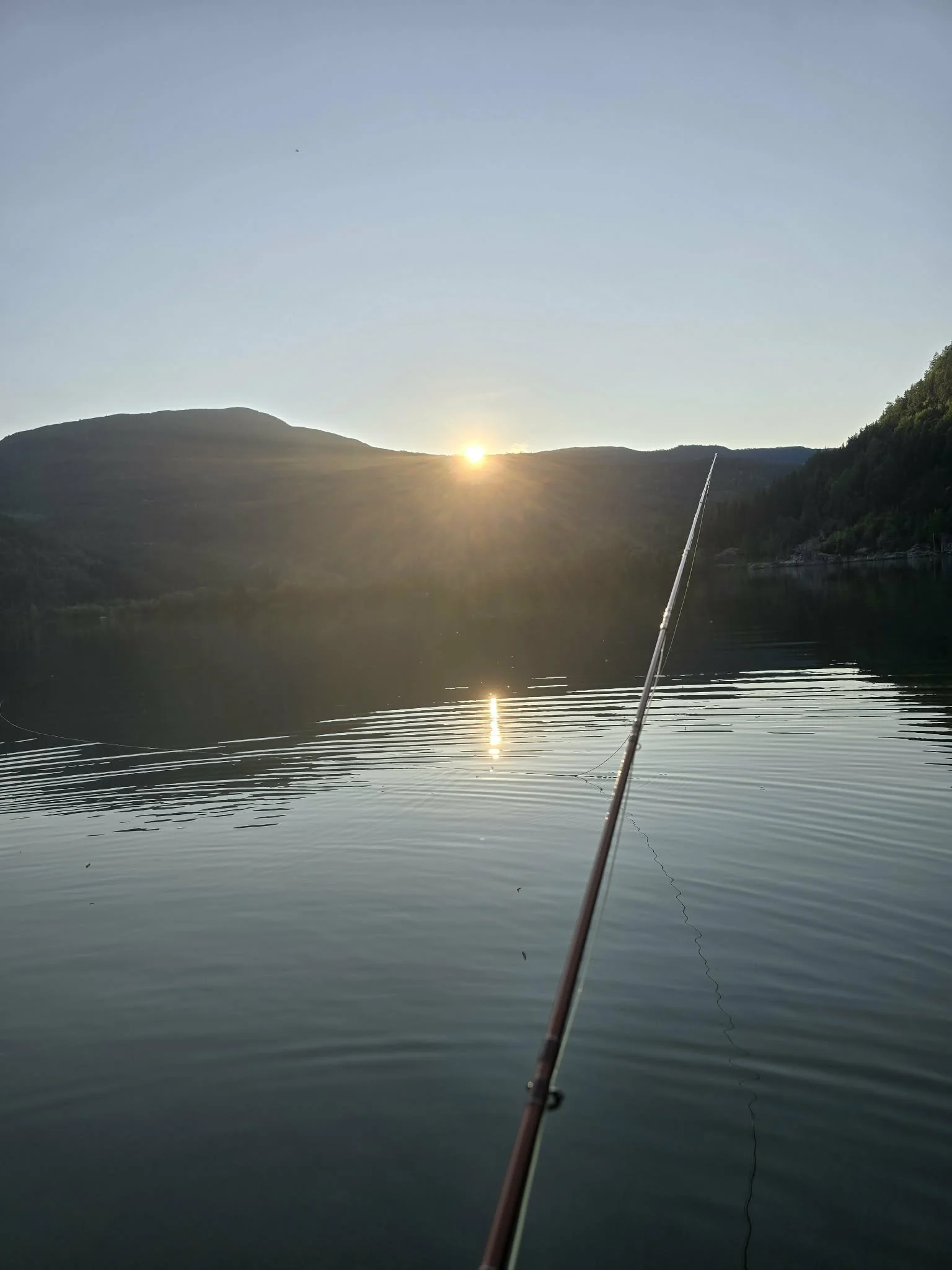 Fiskestang i vann med solnedgang bak fjell og stillestående vann.