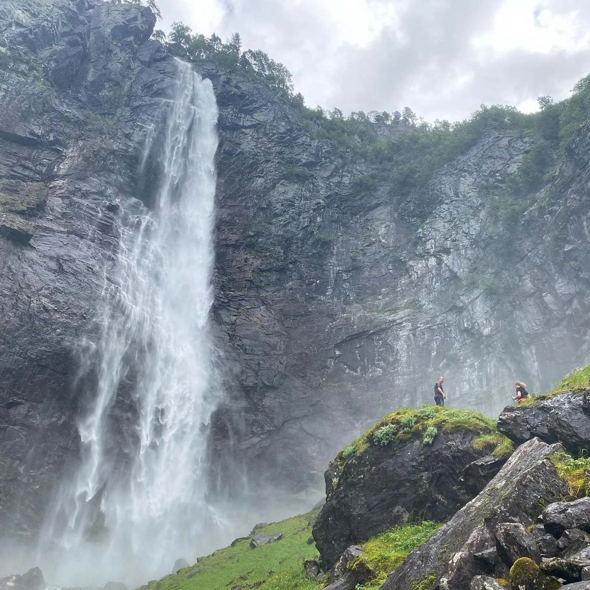 To personer står på grønne steiner ved en foss i en fjellrik natur