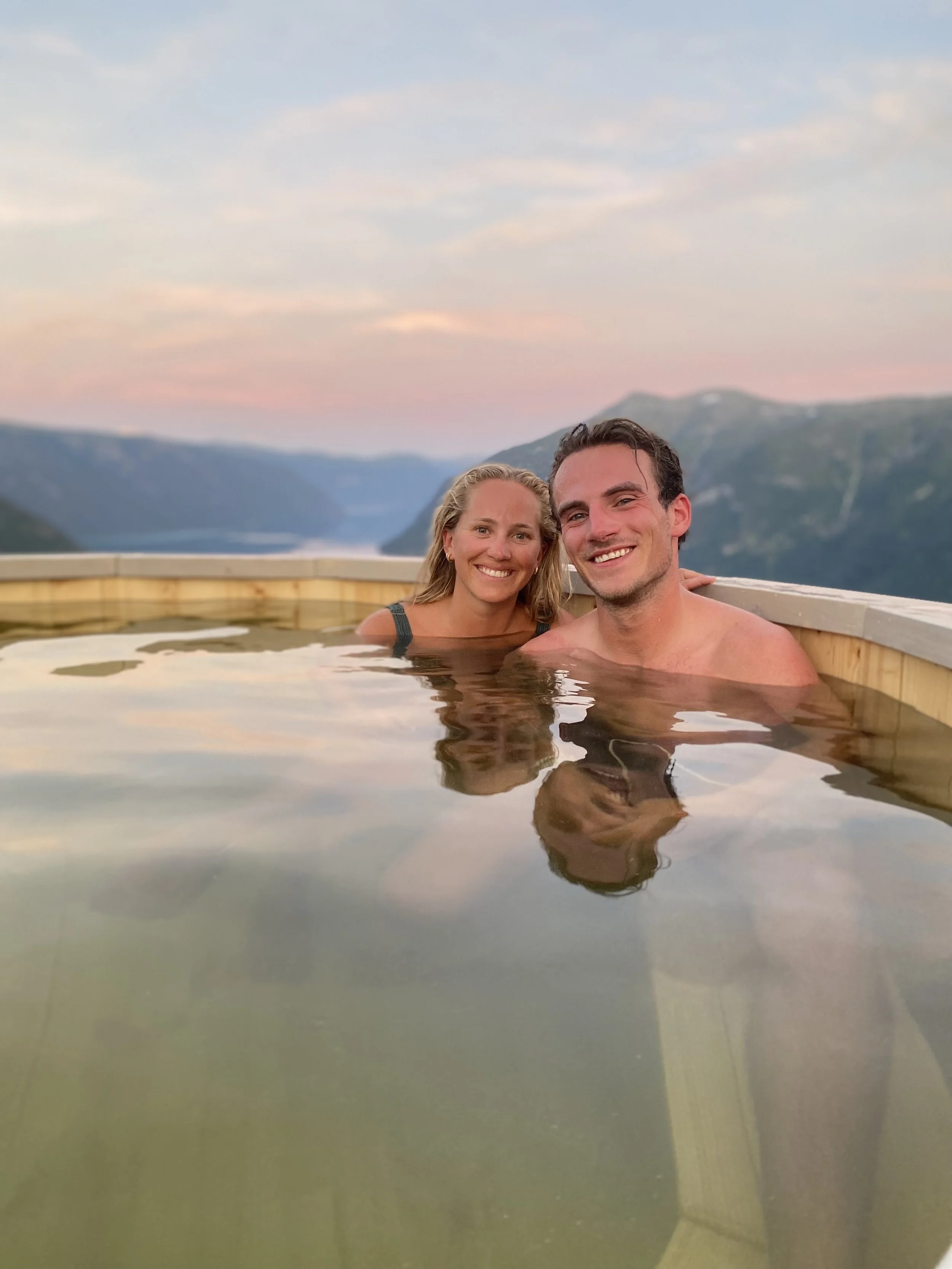 To personer nyter et utendørs varmtbad med fjell og solnedgang i bakgrunnen.